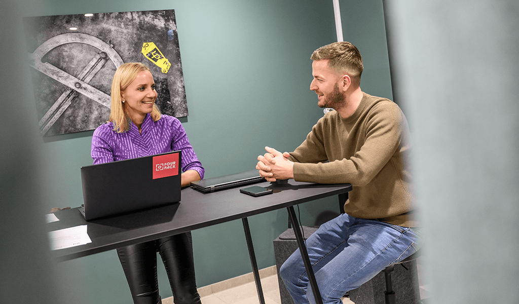 Two employees talking at a table in the office, with a FOUR PACK laptop.