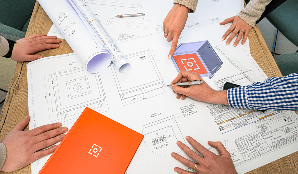 Hände mehrerer Personen über Bauplänen mit einer orangefarbenen FOUR PACK Mappe.