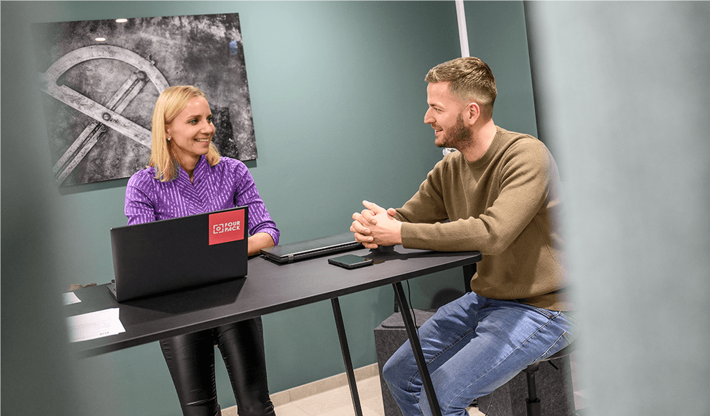 Zwei Mitarbeiter im Gespräch an einem Tisch im Büro, mit FOUR PACK Laptop.
